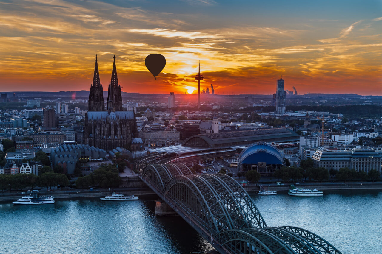 Luftaufnahme von Köln bei Sonnenuntergang von der Deuzer Rheinseite mit Blick Richtung Stadt-Zentrum. aufgenommen. Zu sehen der Reihn die Hohenzollernbrücke, der Dom und der Fernsehturm. Zwischen diesen beiden schwebt ein Fesselballon.