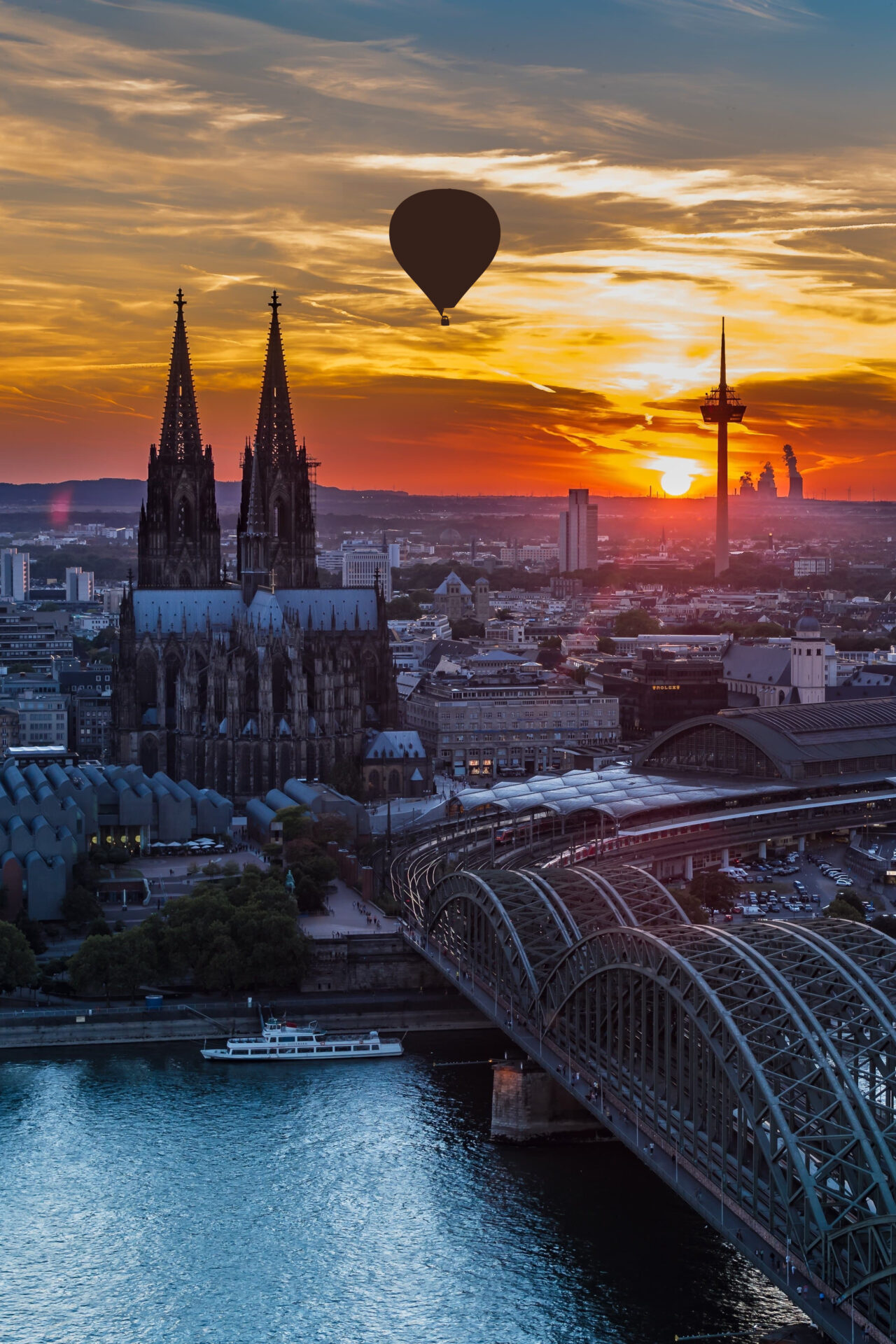 Luftaufnahme von Köln bei Sonnenuntergang von der Deuzer Rheinseite mit Blick Richtung Stadt-Zentrum. aufgenommen. Zu sehen der Reihn die Hohenzollernbrücke, der Dom und der Fernsehturm. Zwischen diesen beiden schwebt ein Fesselballon.